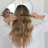 Person combing their long, wavy hair using a VESITIA comb in front of a mirror.