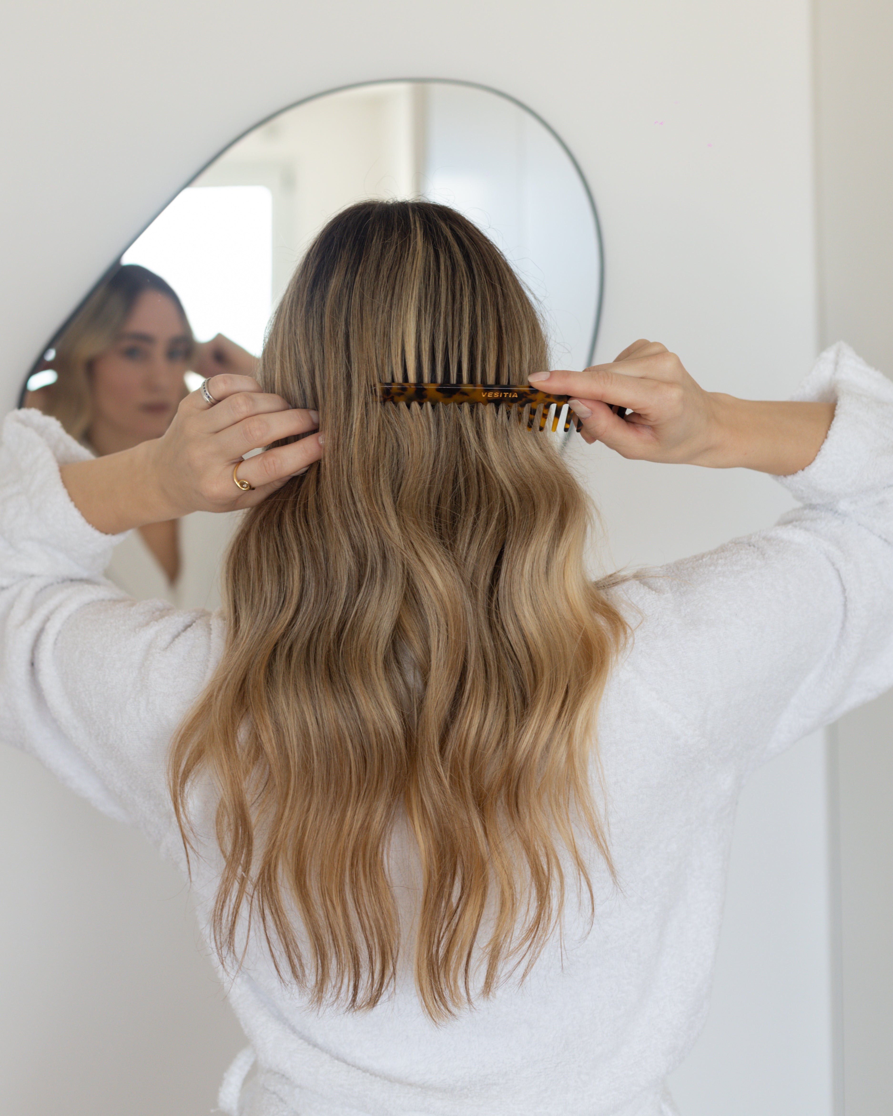 Person combing their long, wavy hair using a VESITIA comb in front of a mirror.