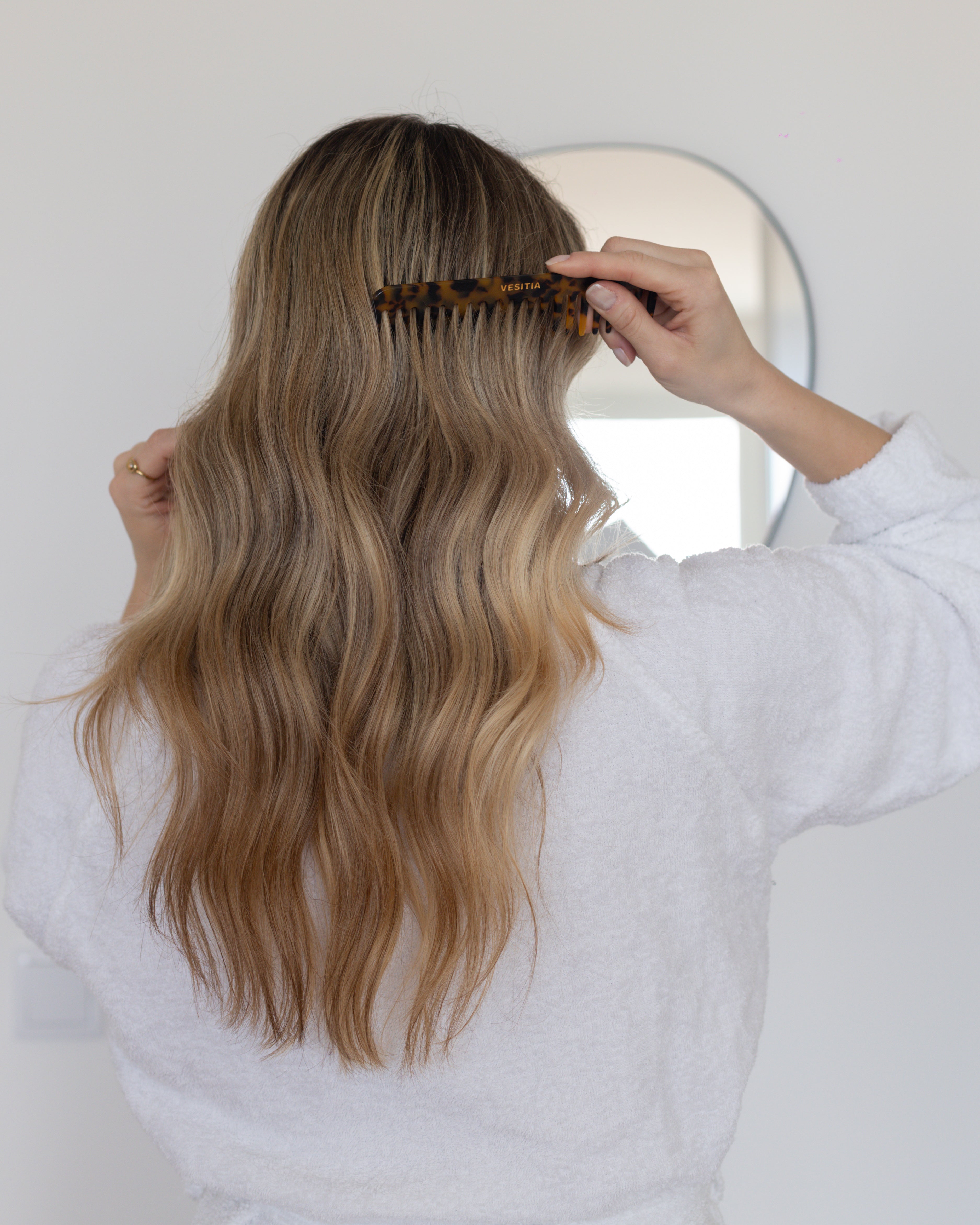 Person combing long, wavy hair with a VESITIA comb in a bathroom setting