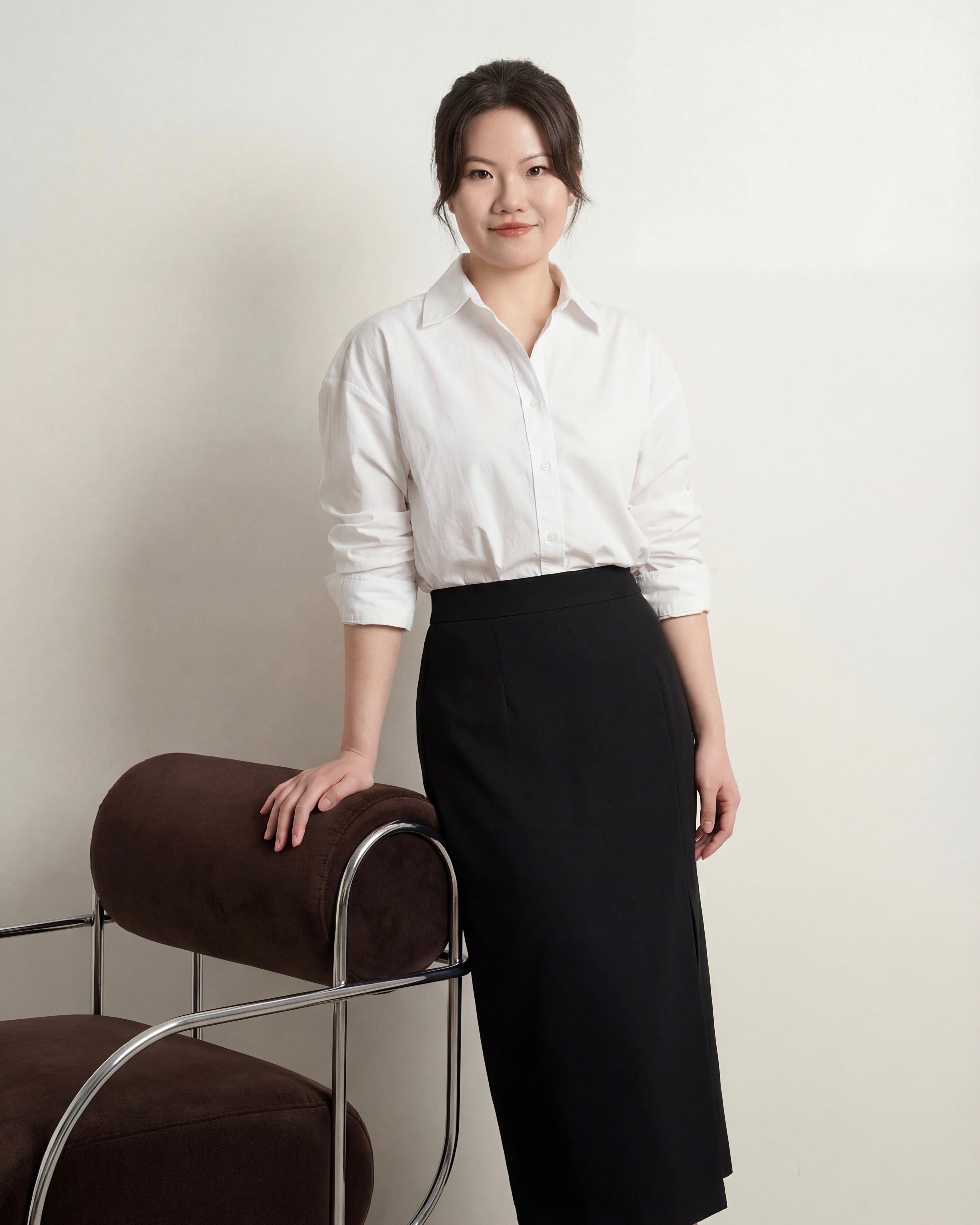 Woman in a white shirt and black skirt standing next to a brown chair against a plain background