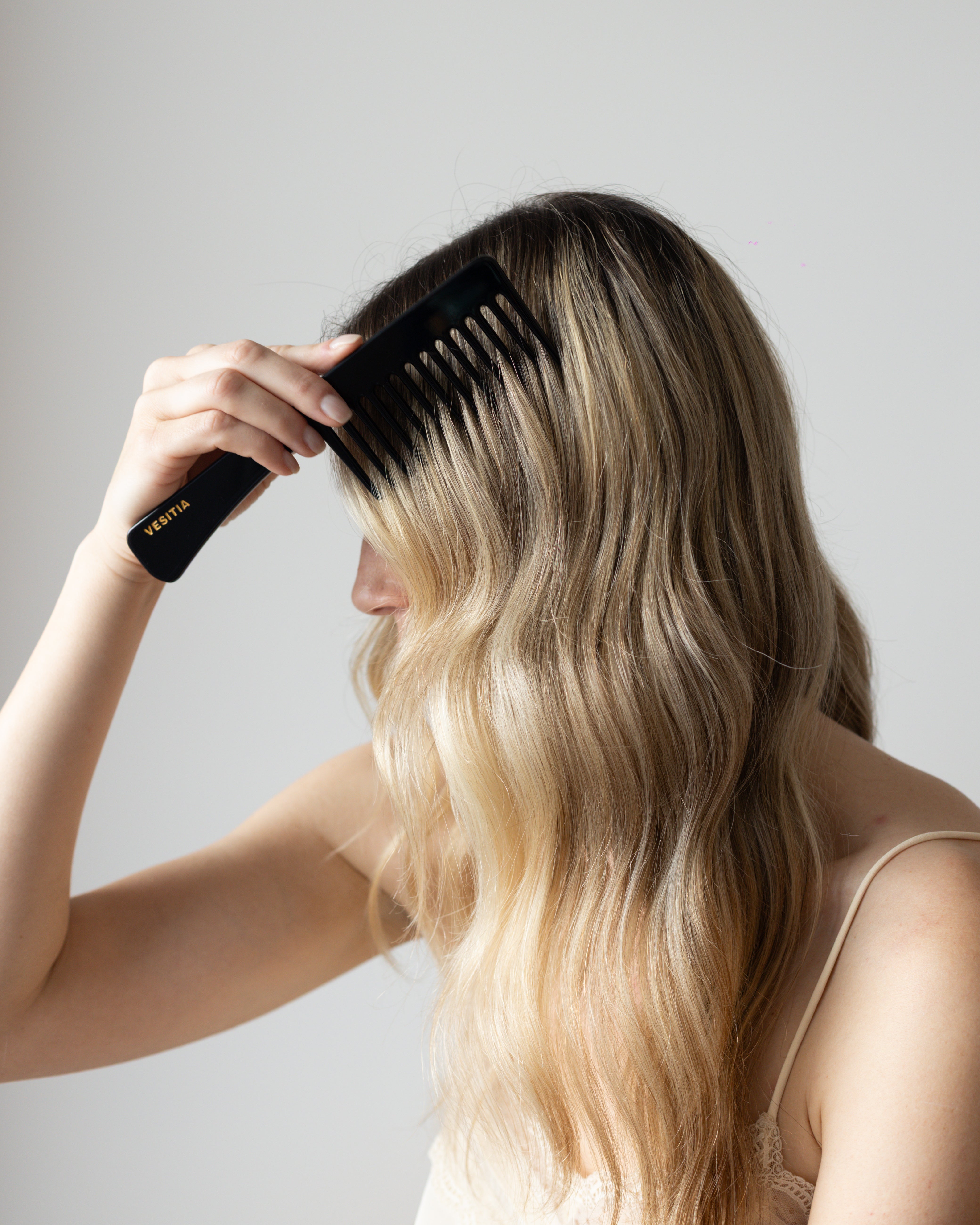 Person with long blonde hair using a black VESITIA comb on a plain background