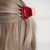 Red hair clip with 'VESITIA' branding on blonde hair against a white background