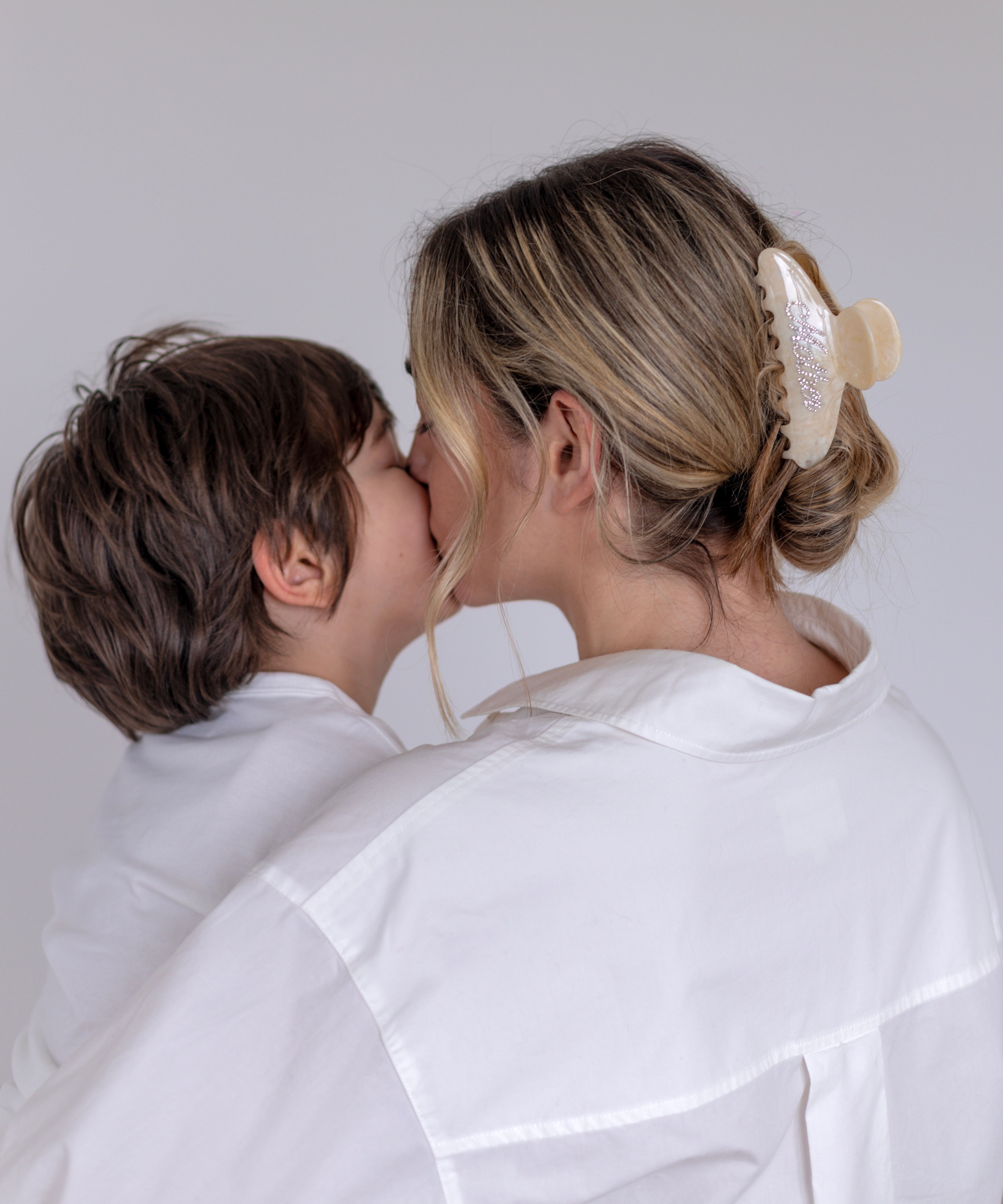 Woman in a white outfit wearing a VESITIA mother claw clip kissing a child on a plain background