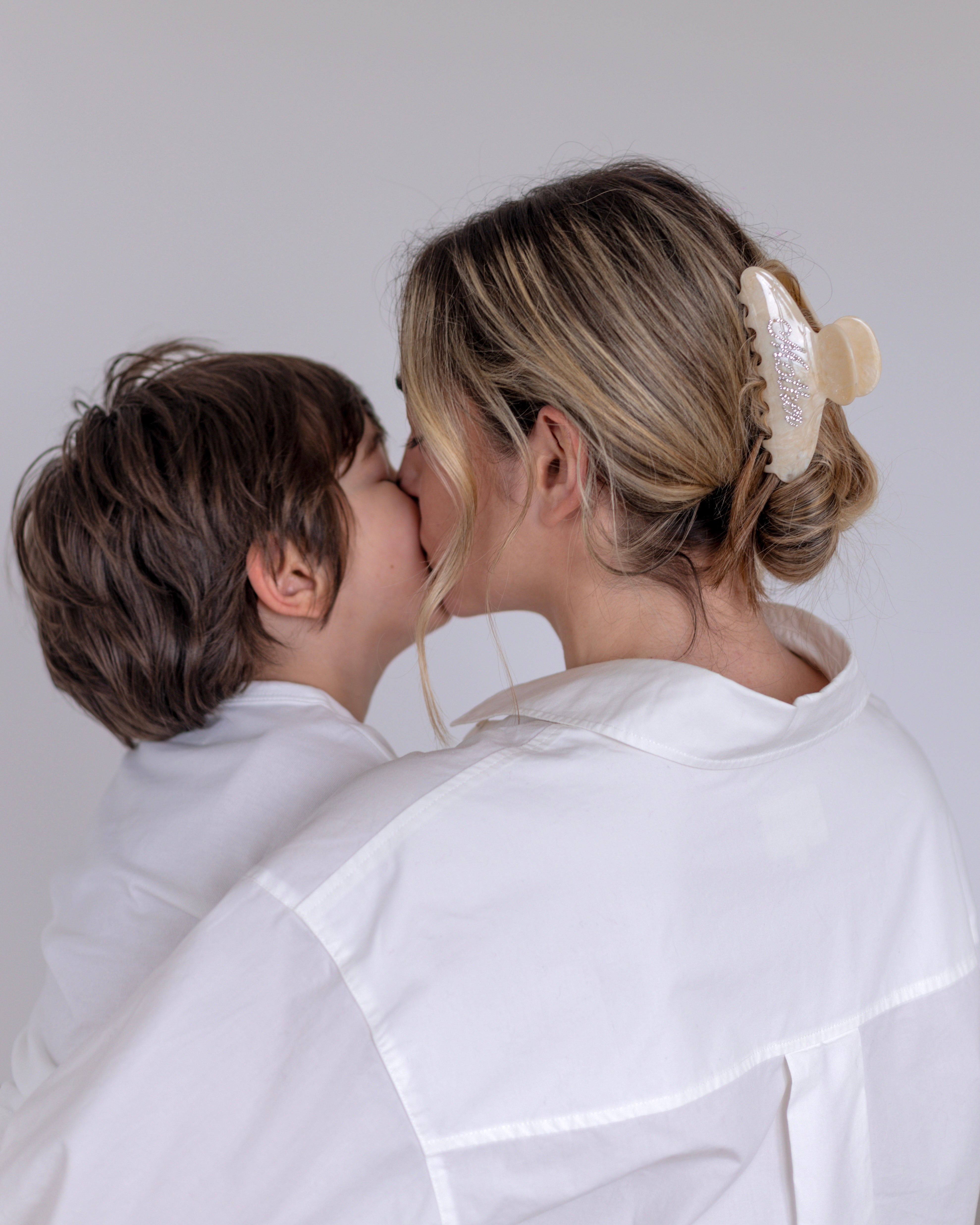 Woman in a white outfit wearing a VESITIA mother claw clip kissing a child on a plain background