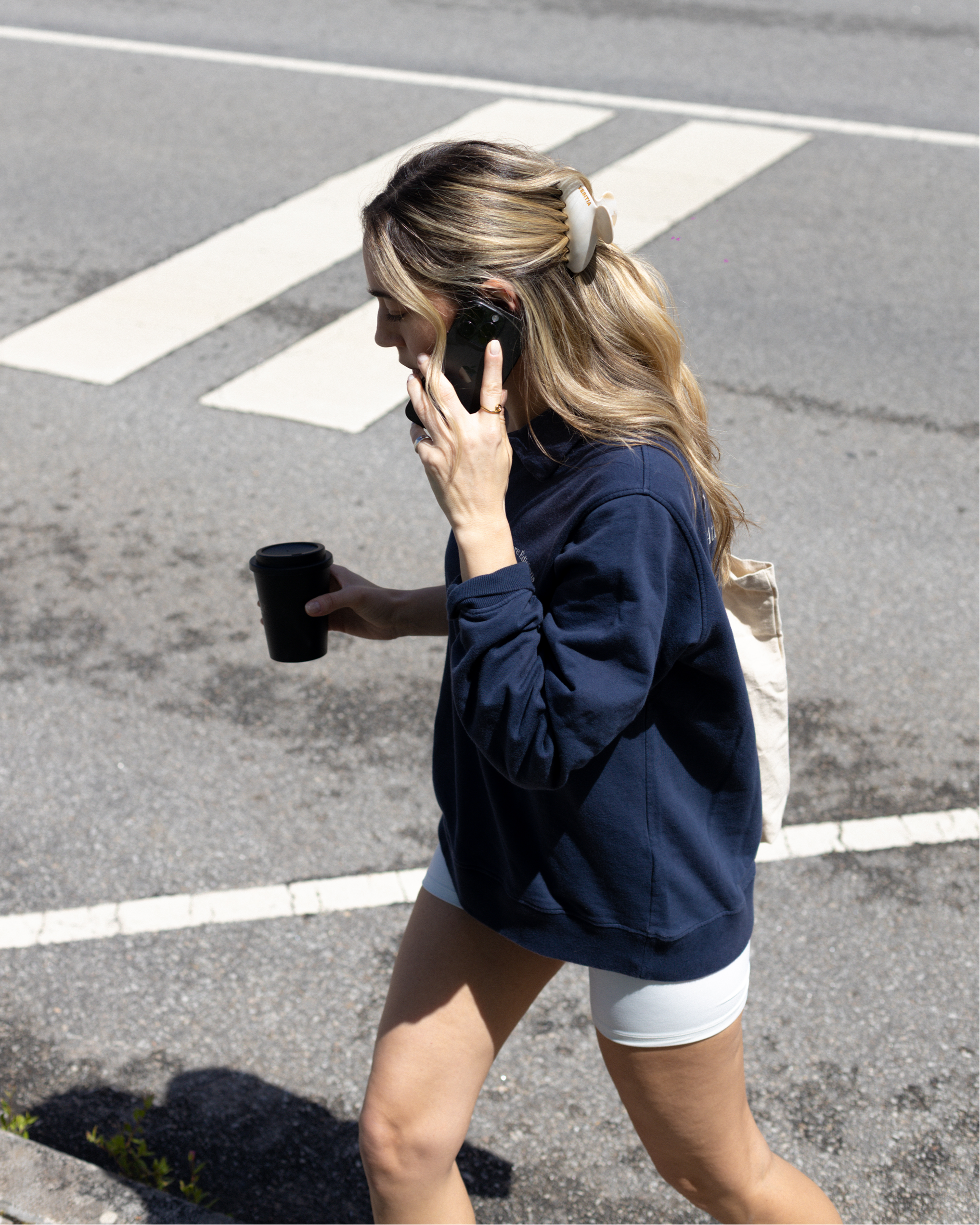 Woman walking on a street, hair styled with a VESITIA claw clip, talking on a phone and holding a coffee cup.