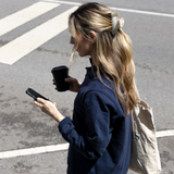 Woman in navy blue sweatshirt wearing a VESITIA claw clip and holding a phone and black cup on a street.