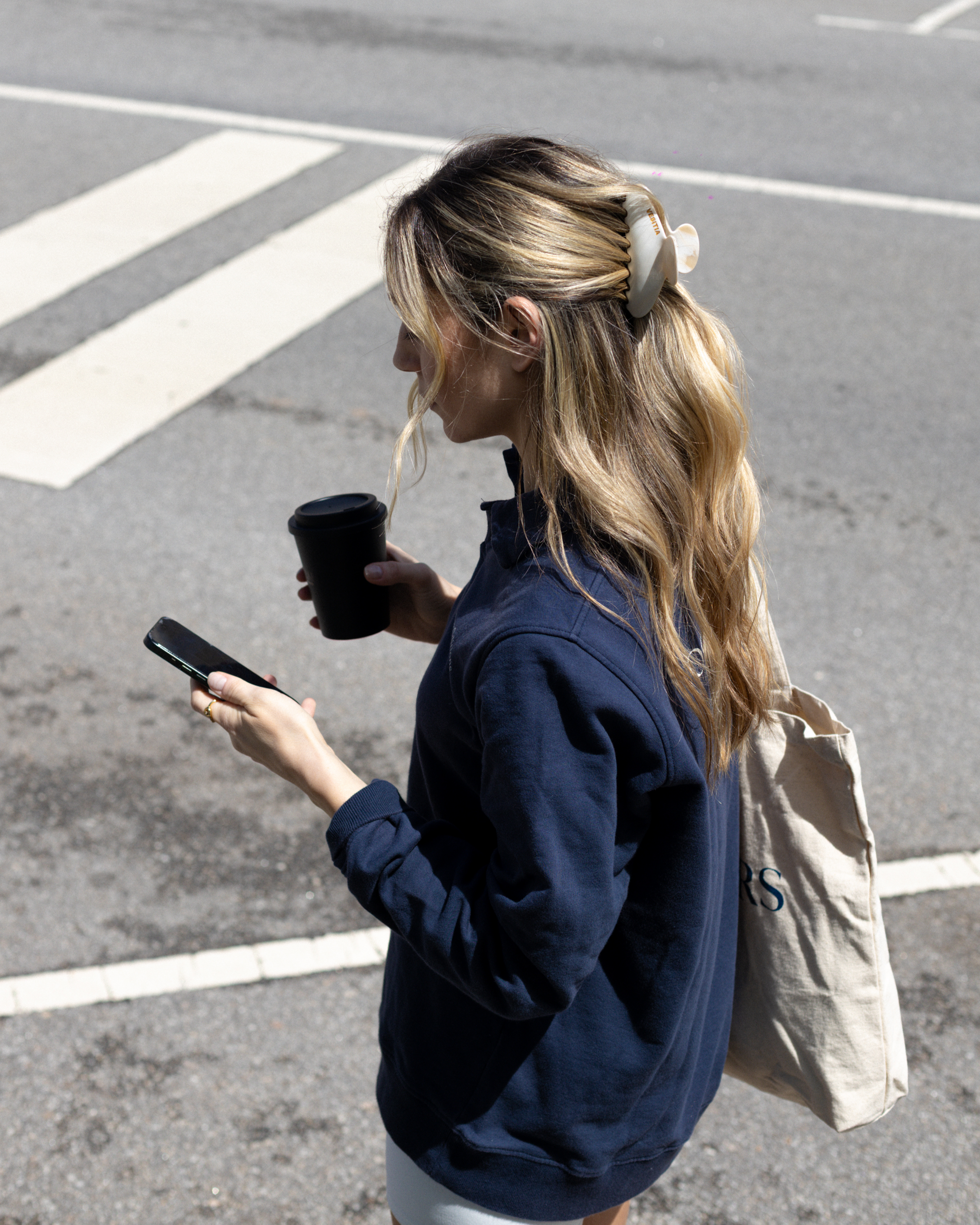 Woman in navy blue sweatshirt wearing a VESITIA claw clip and holding a phone and black cup on a street.