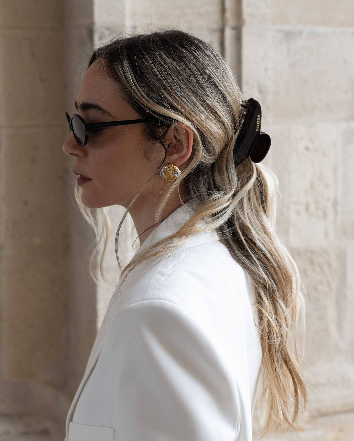 Woman with blonde hair wearing a VESITIA claw clip, asunglasses and a white blazer against a stone wall.