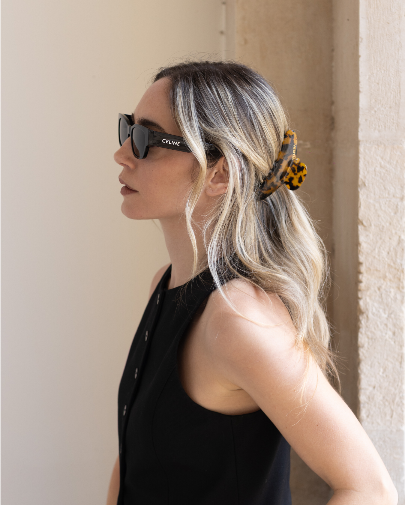 Woman wearing sunglasses with a tortoise VESITIA claw clip against a neutral wall.